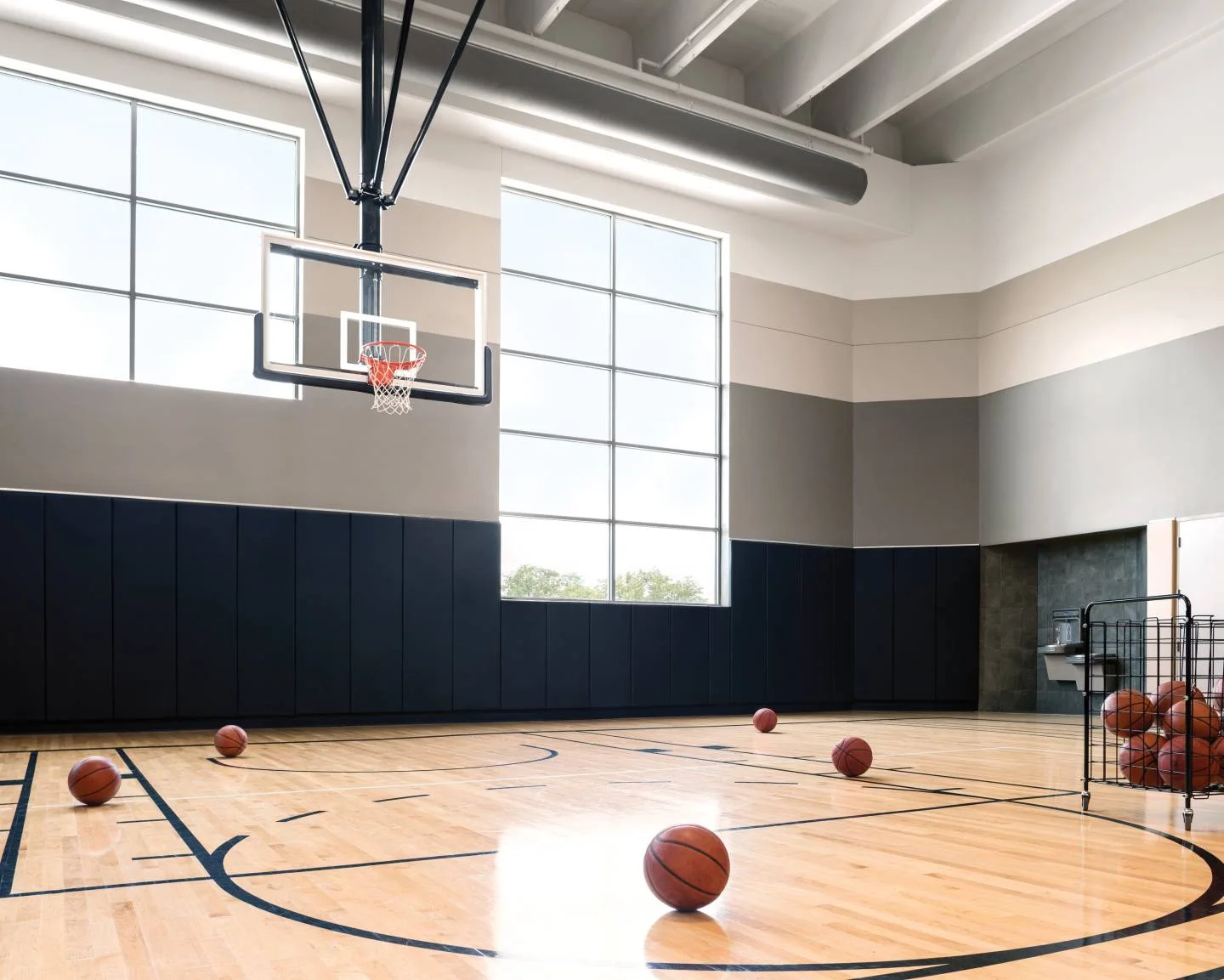 Indoor basketball court with a hoop, large windows and basketballs ready for practice
